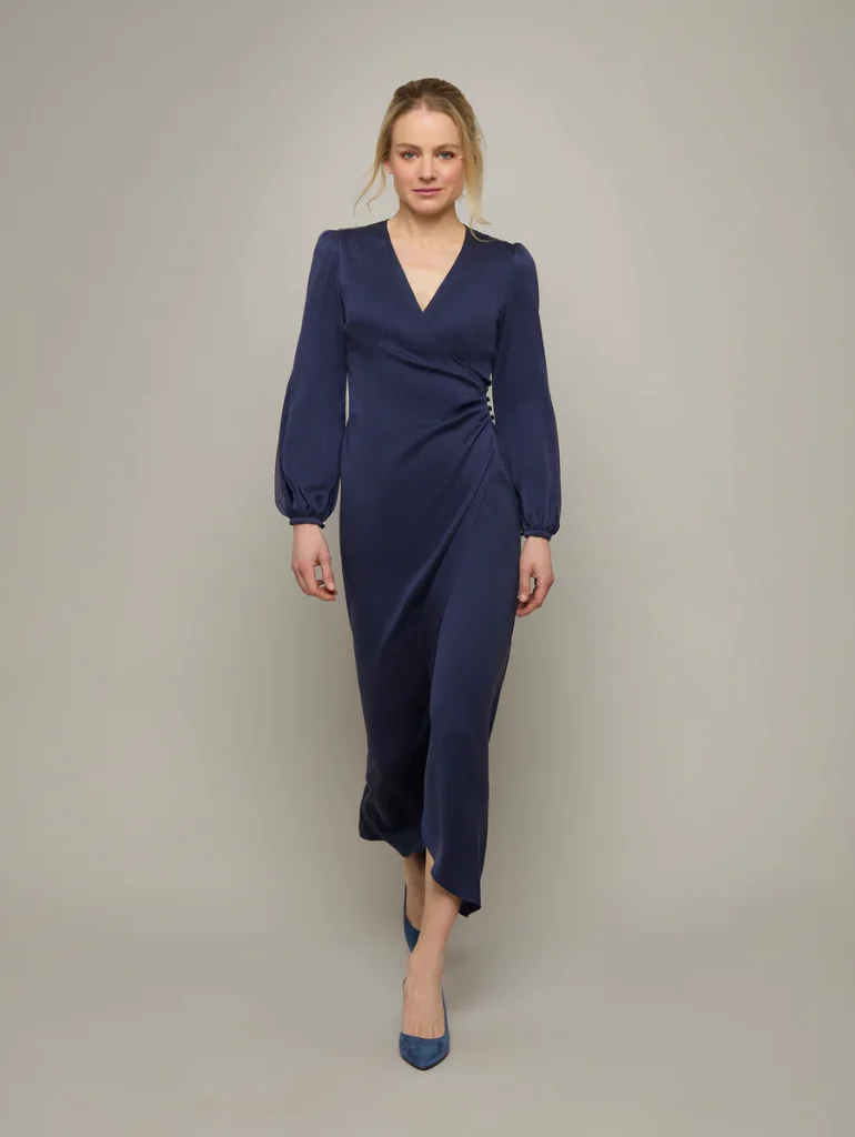 Woman in elegant navy wrap dress and heels, standing confidently against a neutral background