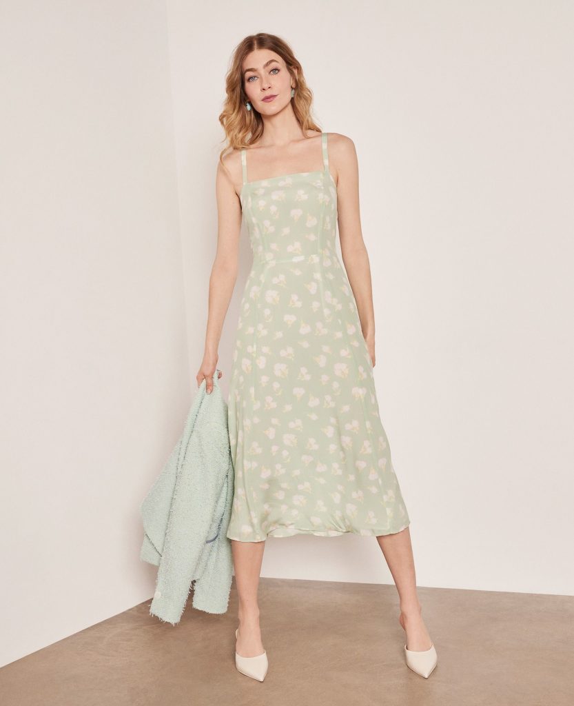 Woman in a green floral dress holding a matching jacket, standing in a minimalist room