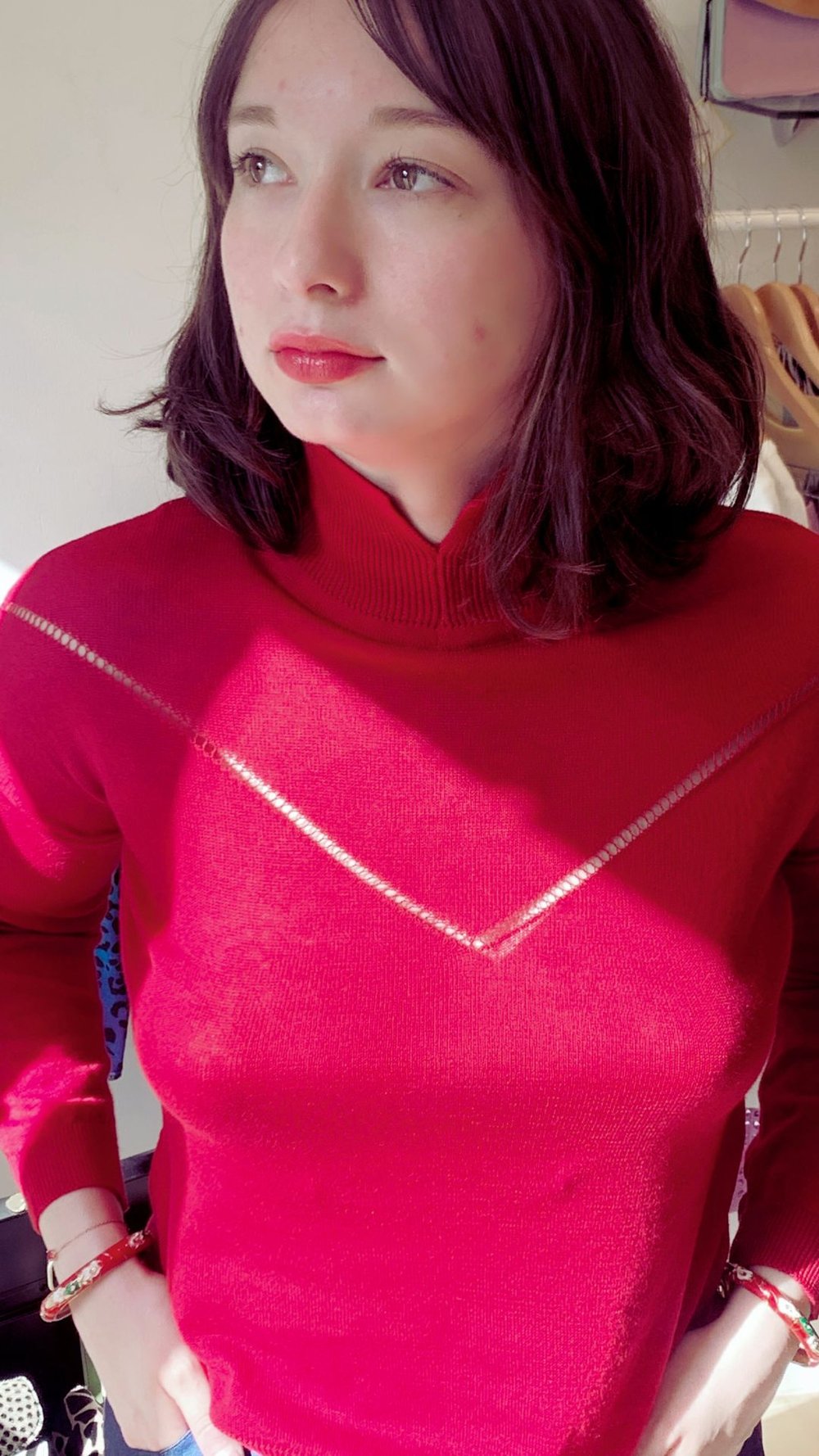 Woman in red sweater with a V pattern looking to the side, standing indoors with clothes in the background