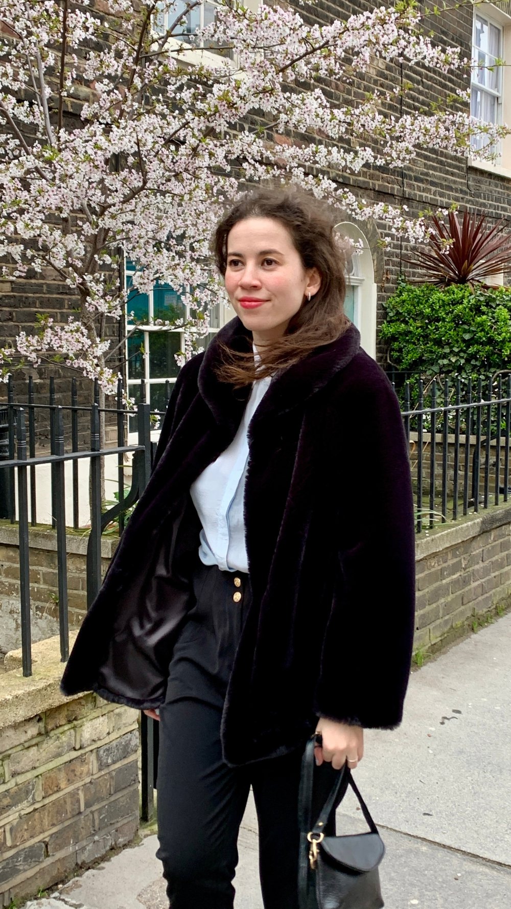 Woman in black coat walking past cherry blossoms and brick building on a spring day.