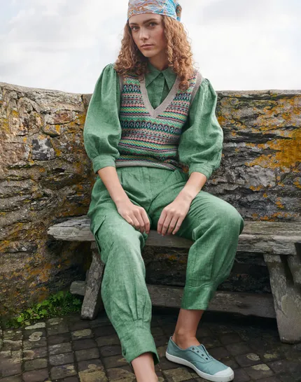 Woman wearing a colorful knit vest, green blouse, and pants, seated on a rustic bench against a stone wall