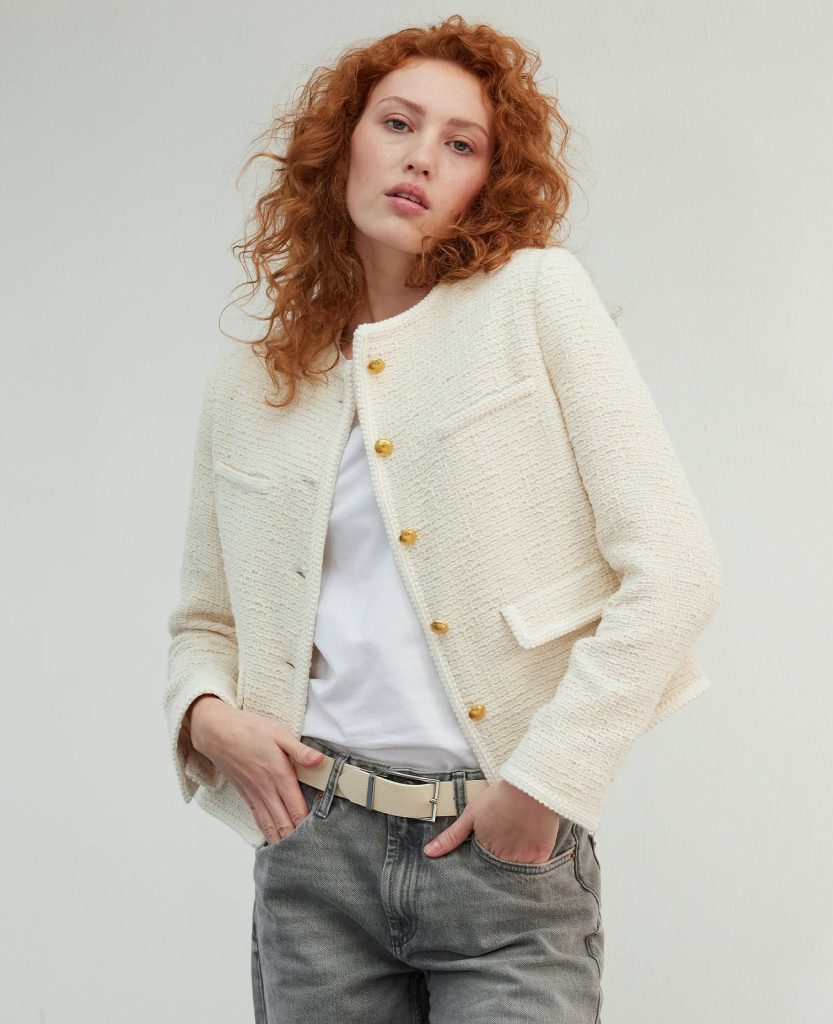 Woman in a stylish cream jacket with gold buttons and gray jeans, hands in pockets, posing confidently
