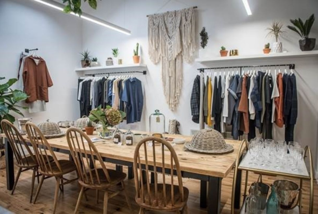 Chic boutique interior with wooden table, woven chairs, hanging clothes, and boho decor against a white wall.