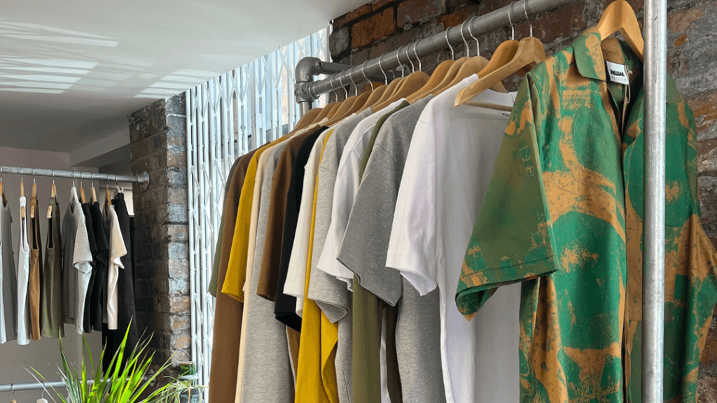 Clothes on hangers in a boutique display various colored shirts against a brick wall.