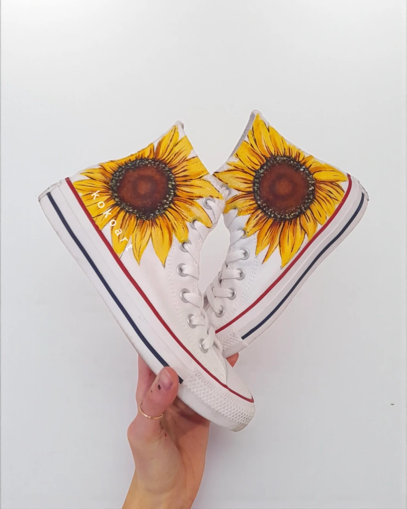 White high-top sneakers with vibrant sunflower art, held by a hand, against a plain background