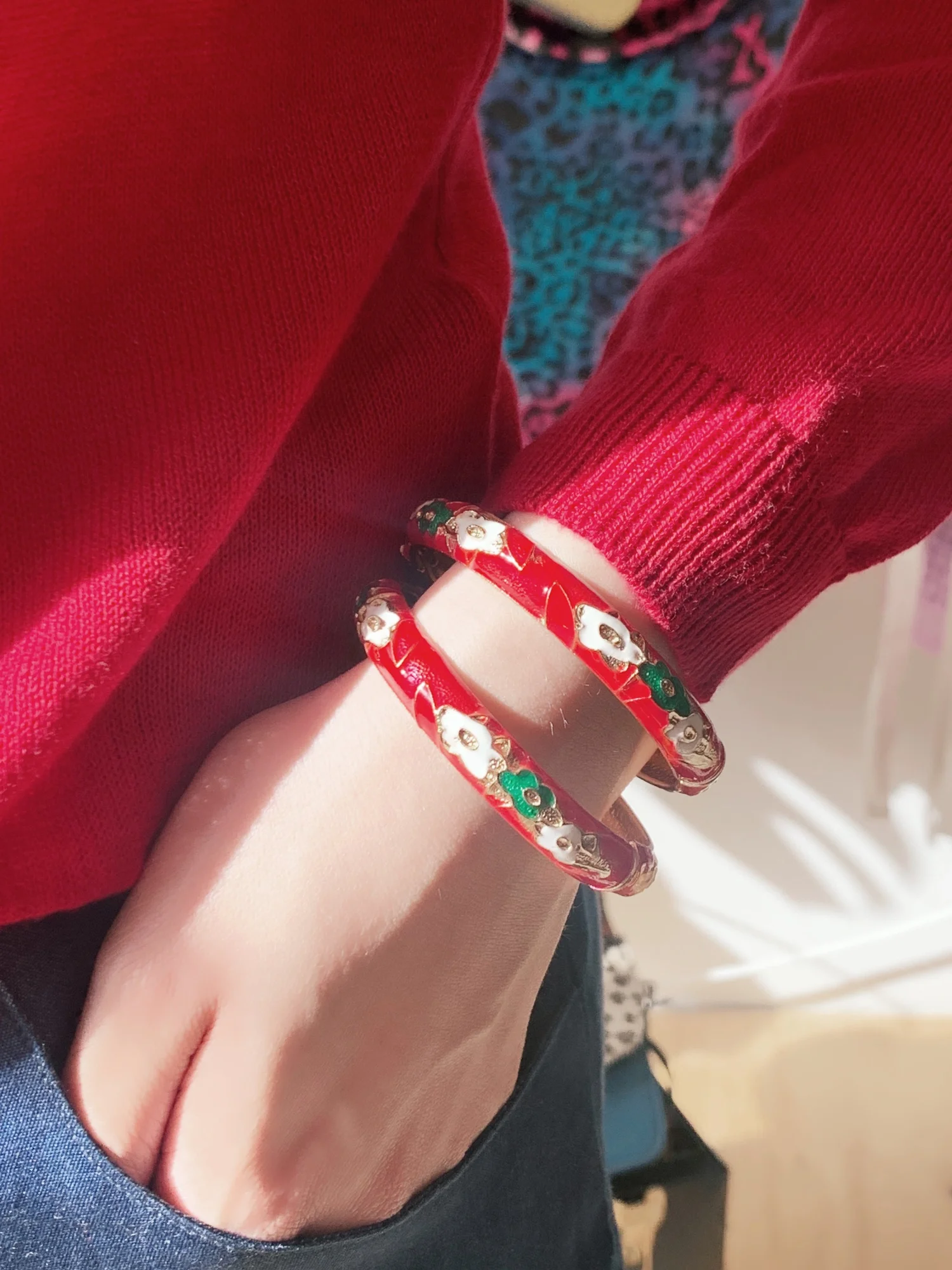 Red enamel bangles with floral design on wrist, paired with red sweater and jeans in bright lighting.