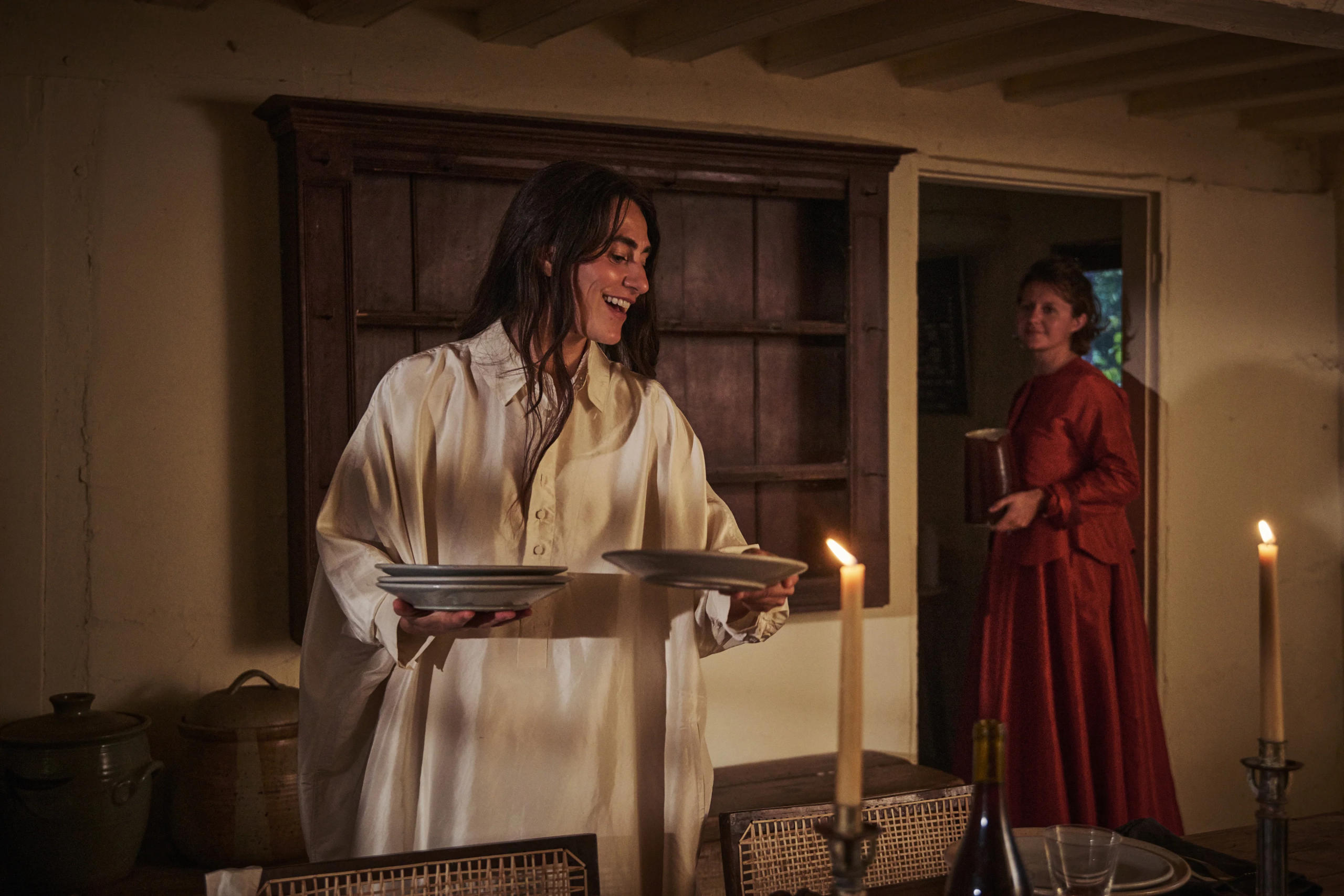 Woman setting dinner table by candlelight in rustic kitchen with another woman in red dress holding a jar.