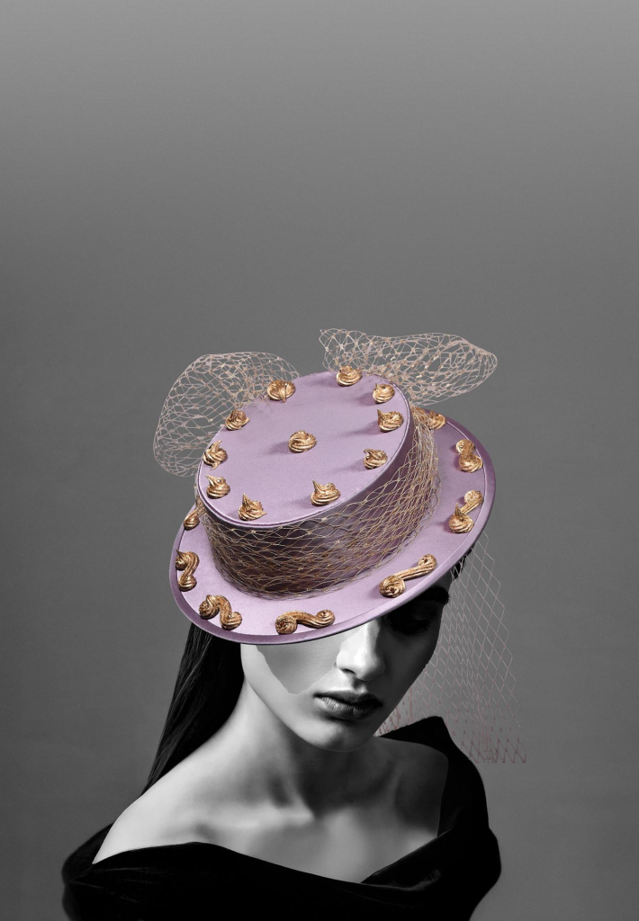 Woman in elegant lavender hat with gold accents and netting, wearing a black off-shoulder dress against gray background