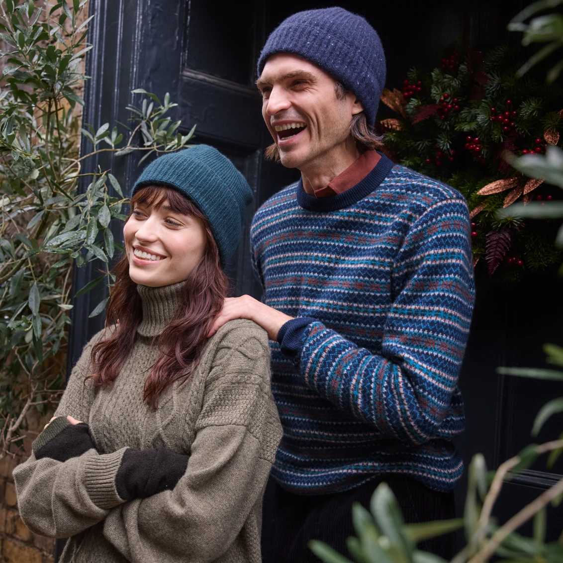 Smiling couple in cosy winter sweaters and beanies, standing by a festive door with a holiday wreath