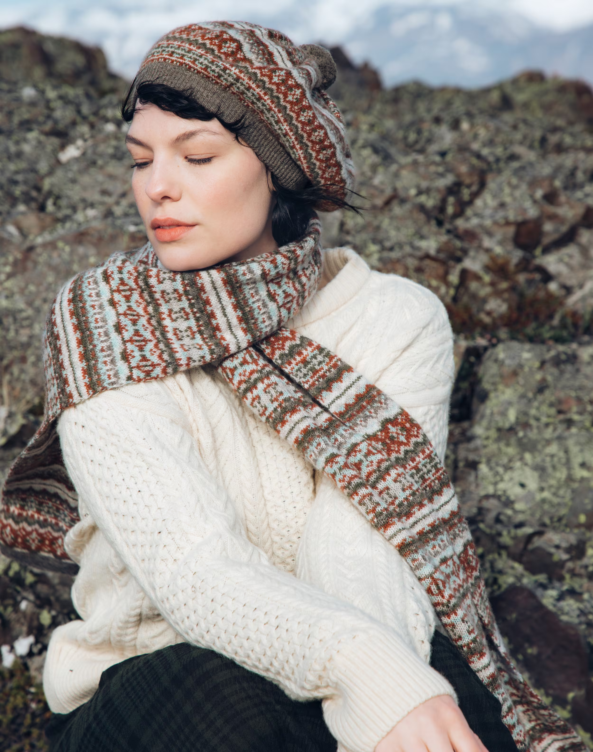 Woman in a cosy knitted sweater, scarf, and hat, sitting outdoors on rocky terrain, enjoying the serene landscape.
