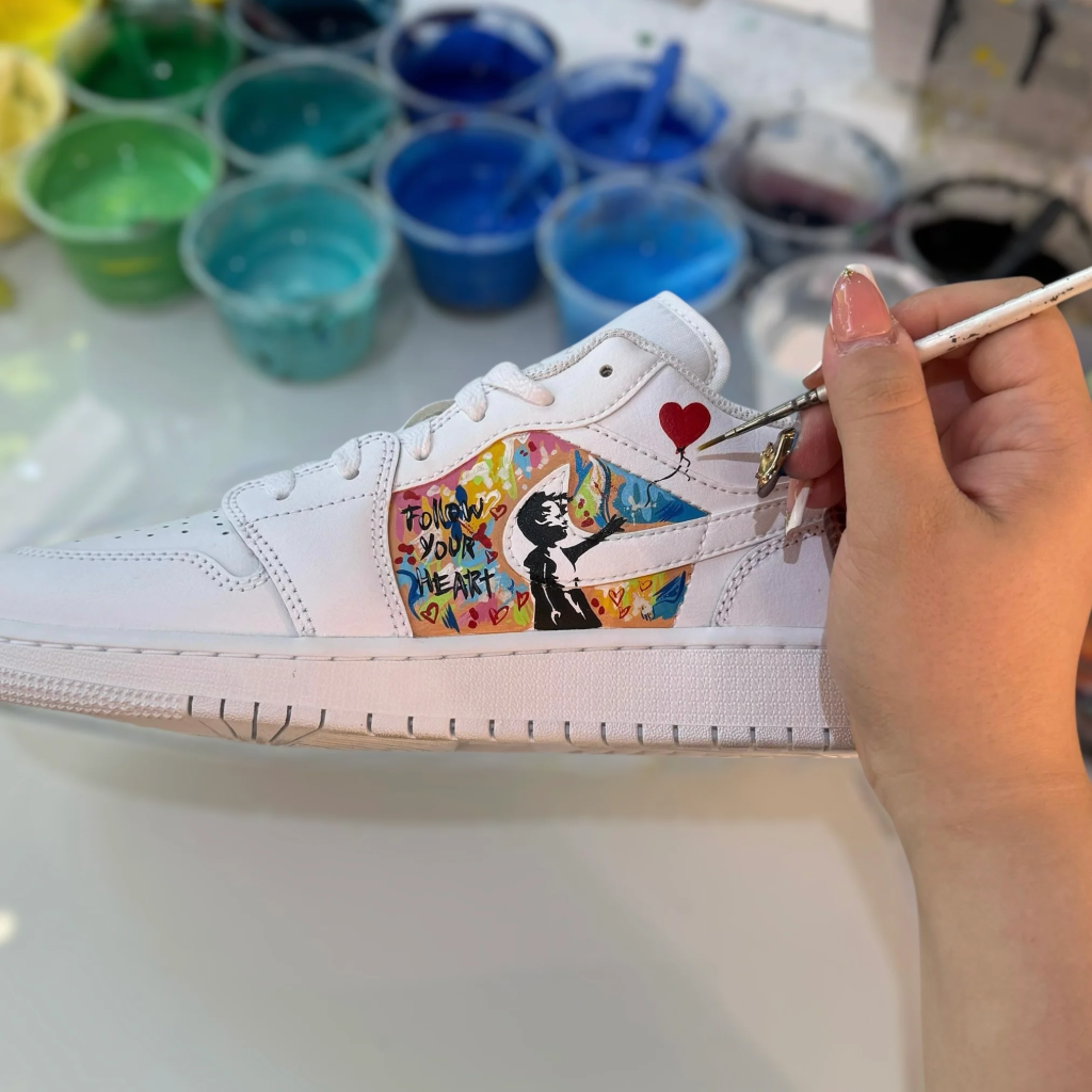 Hand painting a white sneaker with colorful graffiti art and Follow Your Heart text, surrounded by paint cups