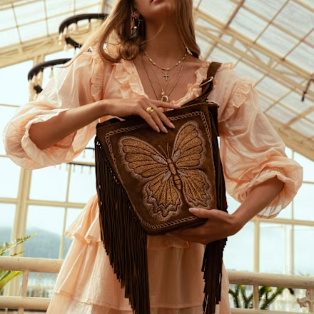 Woman in ruffled dress holding a fringe bag with butterfly design, standing in sunlit room with large windows.