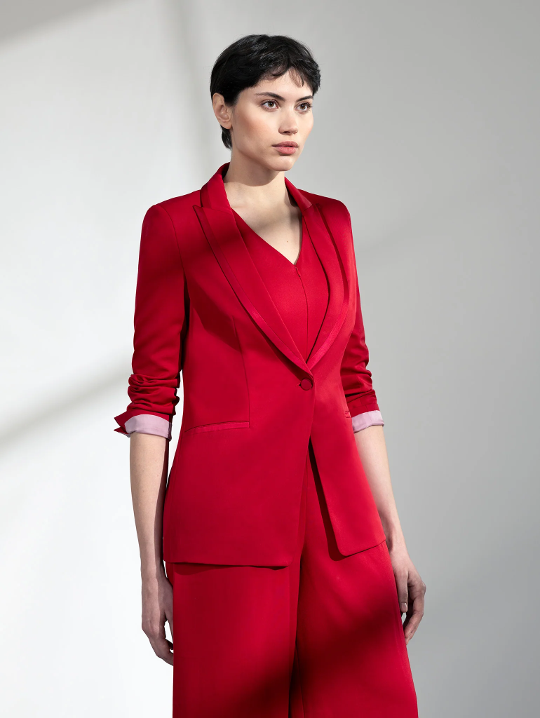 Woman in a stylish red suit poses confidently against a gray backdrop, showcasing modern business fashion