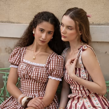 Two women in vintage-style dresses sitting on a bench outdoors, wearing gingham and floral patterns.