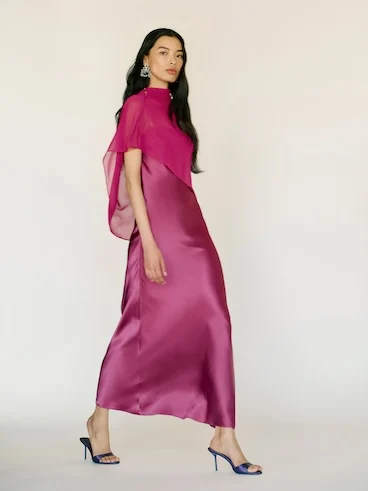 Woman in elegant pink satin dress with sheer overlay, wearing blue heels, walking confidently against a neutral background.