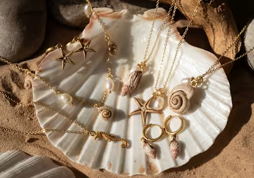 Golden seashell-themed jewelry displayed on a large white shell, featuring necklaces and earrings in a beach setting.