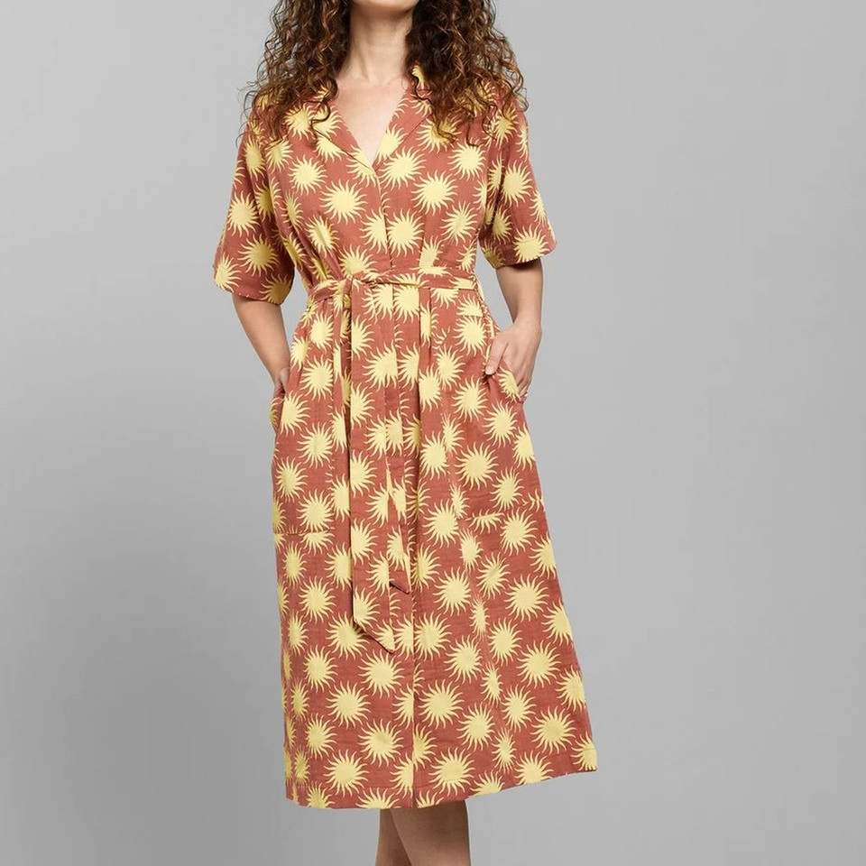 Woman in a patterned, knee-length dress with pockets, featuring yellow sun motifs on a brown background.