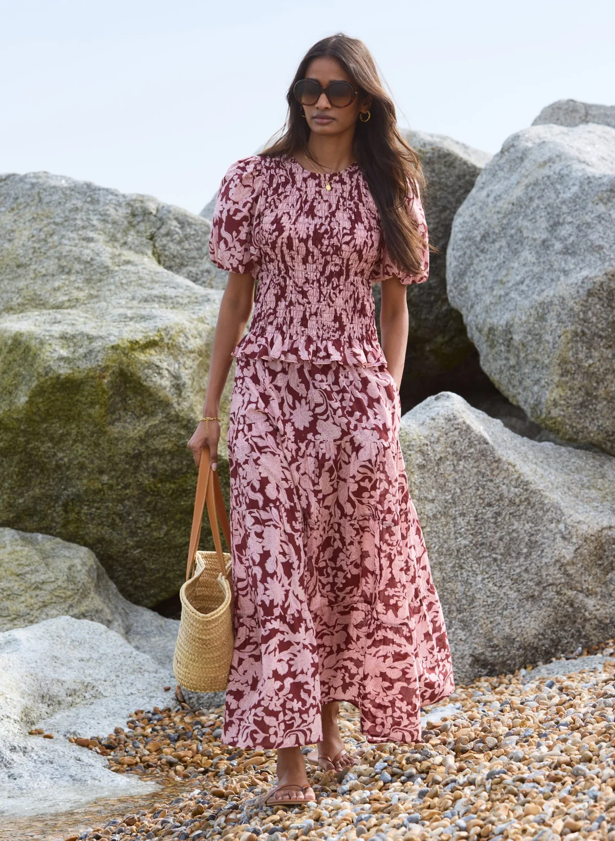 Woman in floral maxi dress and sunglasses walking on a rocky beach carrying a straw tote bag.