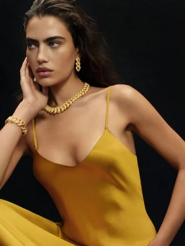 Elegant woman in a golden dress, wearing matching jewelry, posing against a black background, showcasing fashion style.