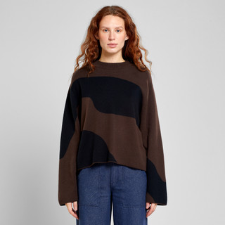 Woman in brown and black abstract sweater with blue jeans, standing against a plain background.