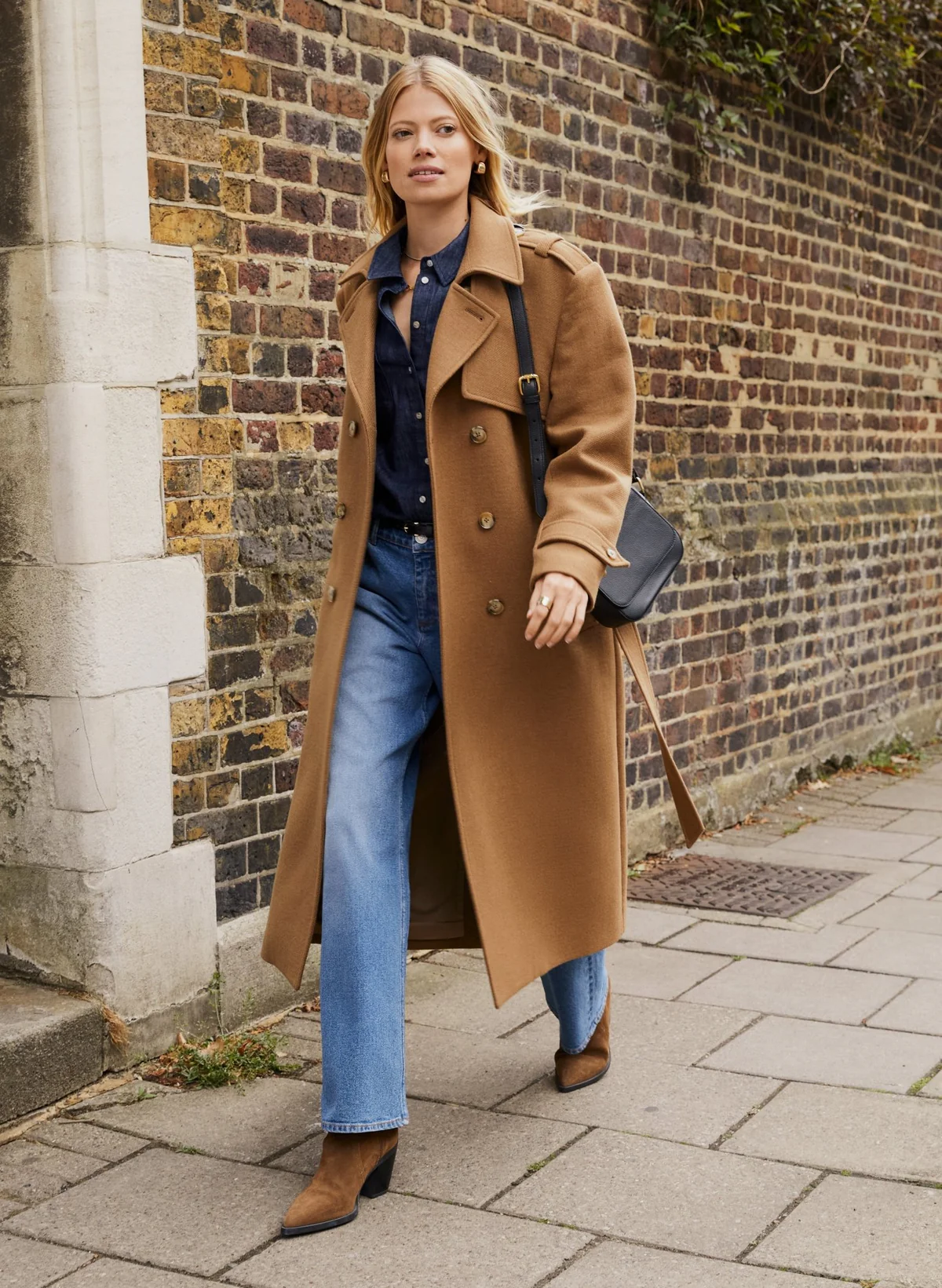 Woman in long brown coat and jeans walking on sidewalk by brick wall.