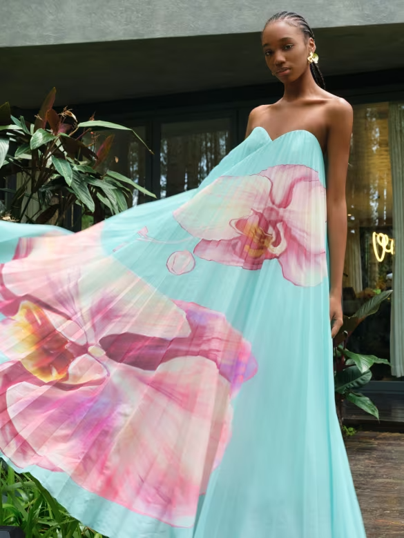 Model in a flowing turquoise dress with large pink floral print, standing outdoors near greenery.