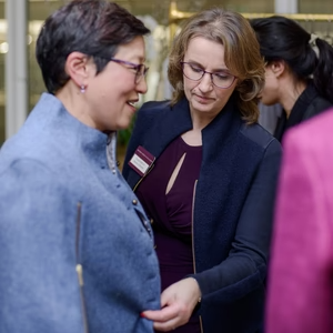 Two women engaging in conversation at a networking event, wearing business attire and glasses.