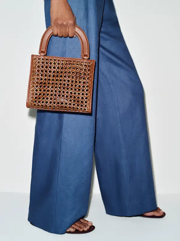 A person holding a stylish brown basket handbag, wearing wide-leg blue trousers and sandals. Fashion accessory focus.